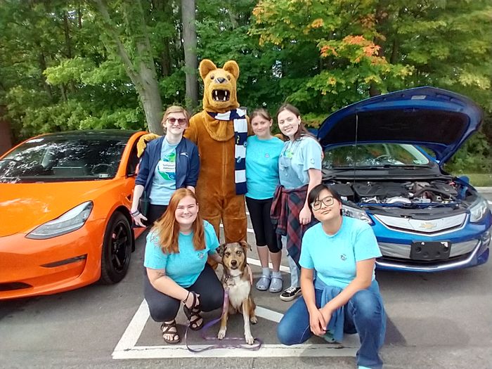 Five college students, Nittany Lion mascot, and dog on leash, near electric cars with trees in background