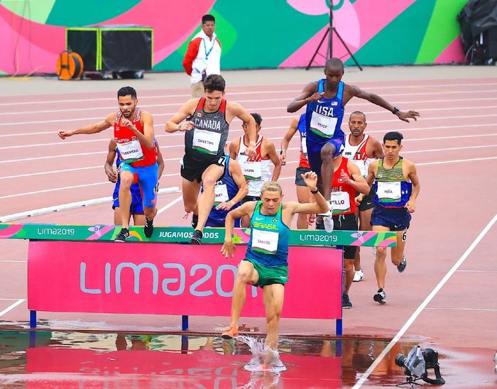 Runners jump over a hurdle and into water during a steeplechase race.