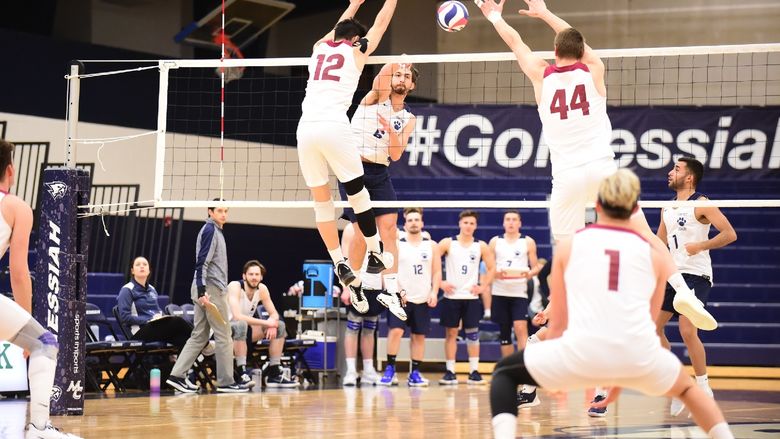 A Penn State Behrend volleyball player jumps to spike the ball over two blocking opponents.