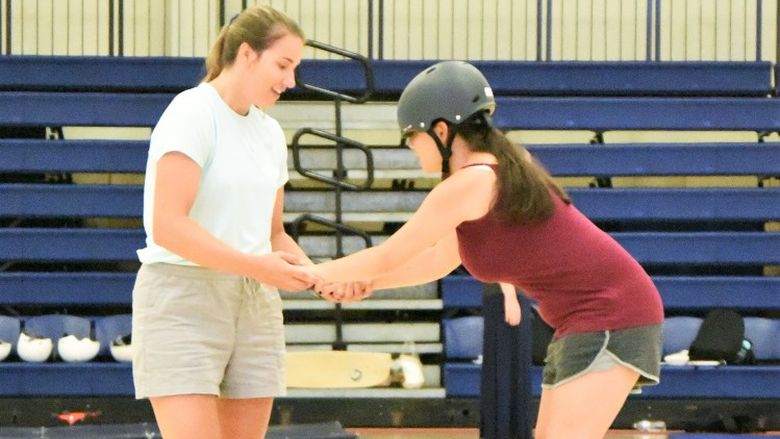 An instructor teaches a blind athlete how to ride a longboard in a gymnasium at Penn State Behrend.