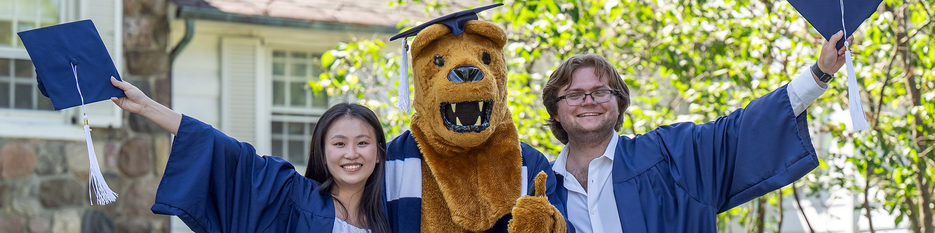 Two graduates in gowns pose with the lion mascot wearing a graduation cap