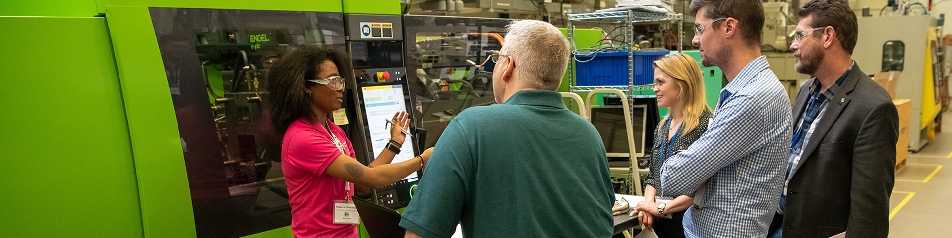 A group of adult students gather around an instructor explaining the operation of a green industrial plastic molding machine. 