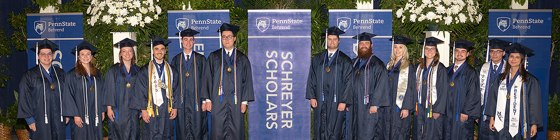 Thirteen Schreyer Honors College graduates in academic gowns and caps stand near banner.