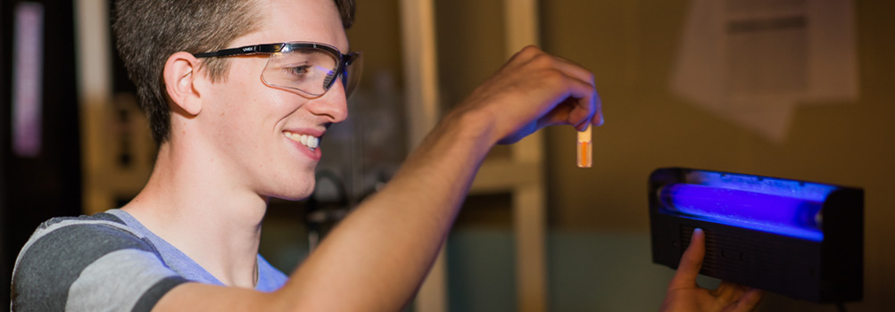 A Penn State Behrend physics student holds a glowing orange vial of liquid in front of a blue light.