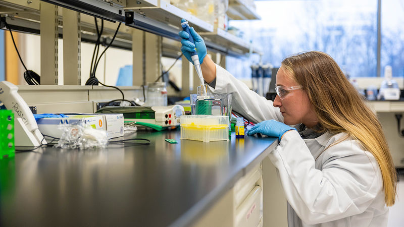 A female student researcher in a lab coat and safety goggles uses a pipette to work with samples in a laboratory setting.