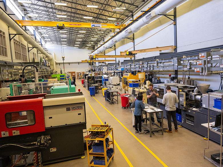 Behrend's large college plastics lab with industrial machinery, including injection molding machines and overhead cranes, where students and an instructor are gathered.