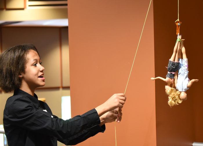 A middle-school student raises a bungee cord that holds two Barbie dolls.