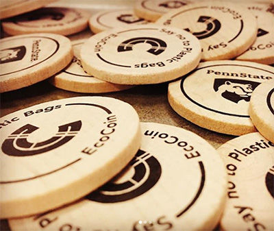 A pile of wooden EcoCoins are scattered. The imprinted text reads, "EcoCoin: Say no to plastic bags."