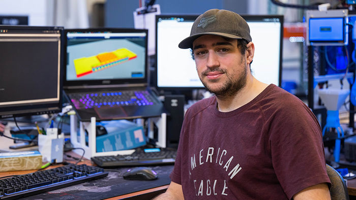 A student wearing a dark baseball cap and a t-shirt sits at a desk with multiple computer screens.