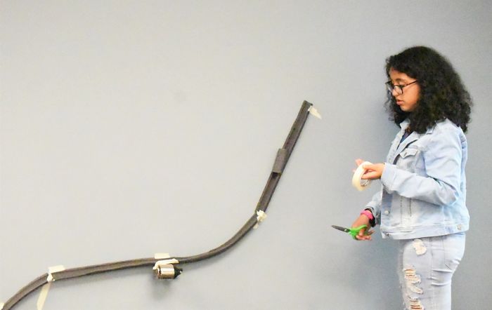 A girl holding tape and scissors stands next to a model roller coaster track, which is hung on the wall with pipe insulators.