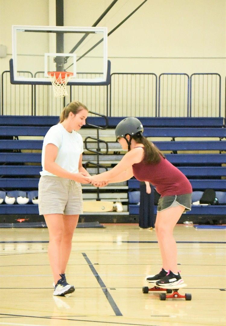 An instructor teaches a blind athlete how to ride a longboard in a gymnasium at Penn State Behrend.
