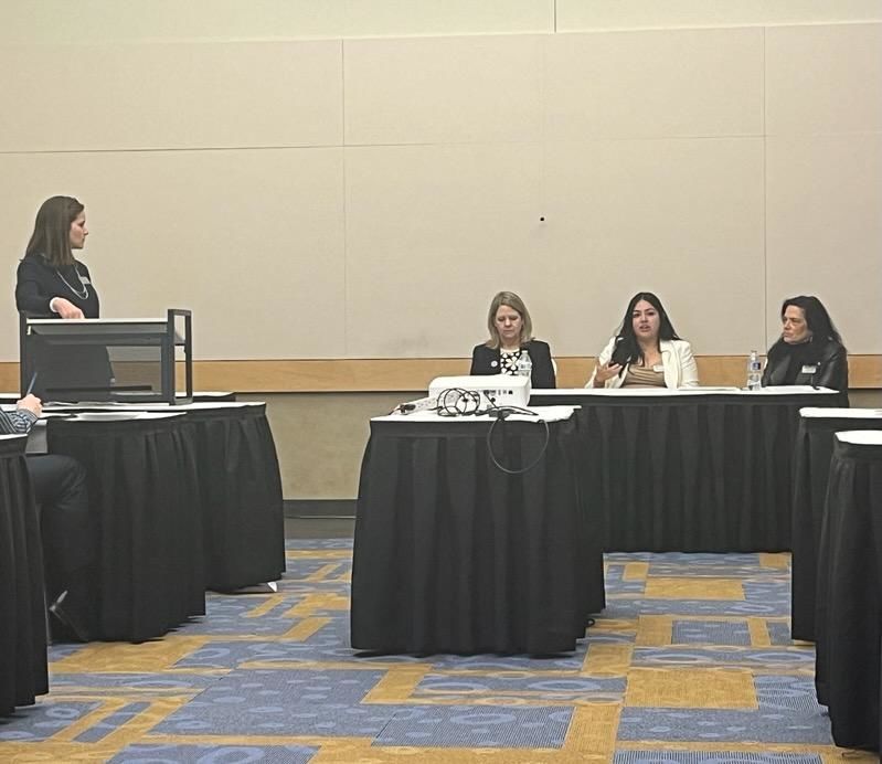 Three women seated behind a table and one at a lecturn. 