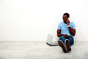 Man sitting and leaning against a walk talking on a phone looking at a computer
