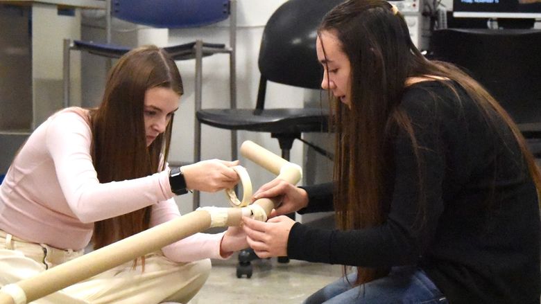 Two female high-school students build a model pipeline with cardboard tubes and masking tape.