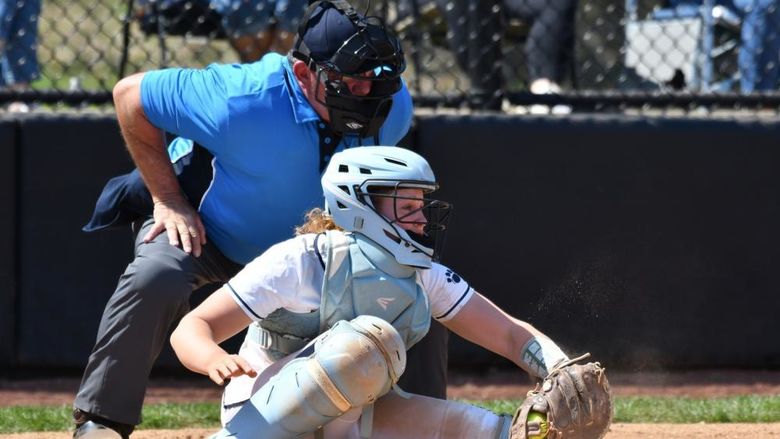 A catcher for the Penn State Behrend softball team grabs the ball as the umpire leans over her shoulder.