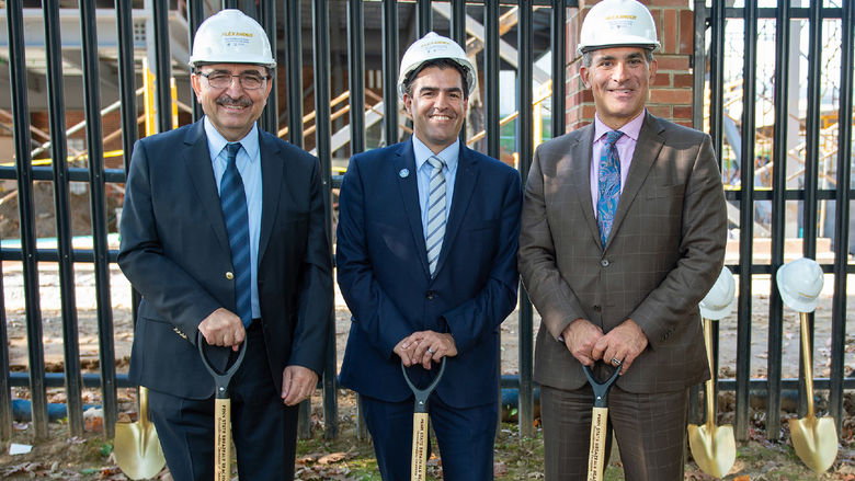Three men wearing suits and hard hats pose with shovels in front of a blank fence.