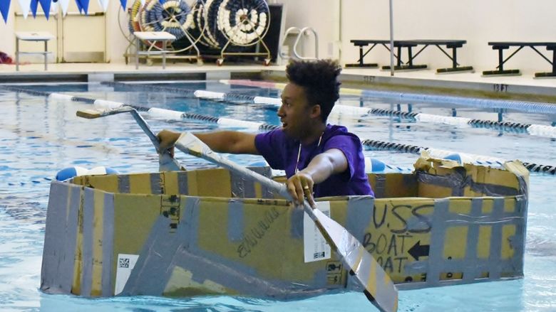 A high school student rows a cardboard boat in a swimming pool