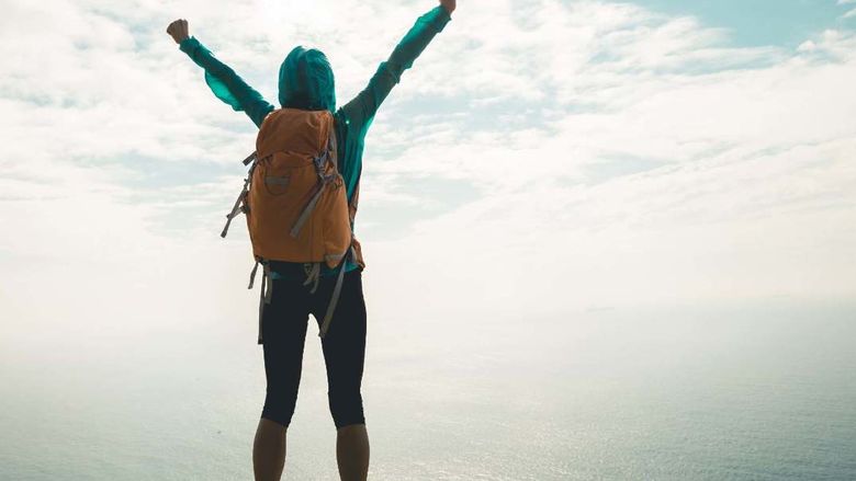A hiker raises his arms at the top of a peak overlooking the ocean.