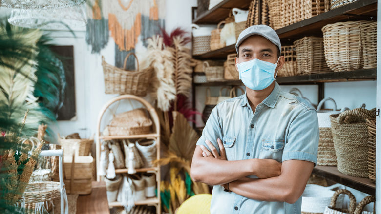 An employee in a hat and facemask poses in a shop stocked with wicker baskets.