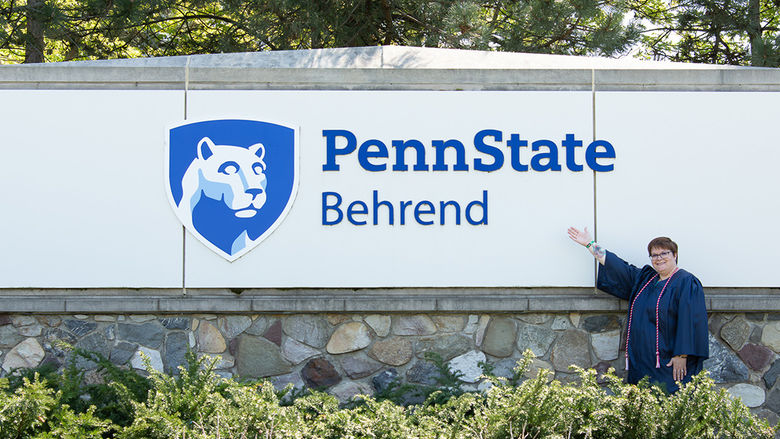 Alice Puzarowski in a graduation cap and gown posing with Penn State Behrend sign