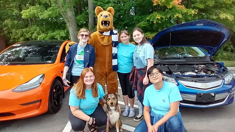Five college students, Nittany Lion mascot, and dog on leash, near electric cars with trees in background