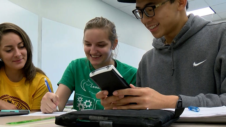 A trio of students work on a project together using pens, rulers, and a calculator.