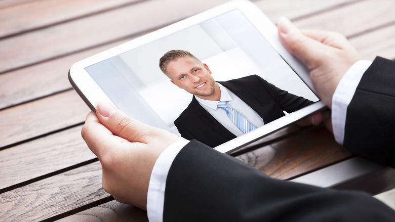 A man in a suit holds a tablet computer that shows another man in a suit.