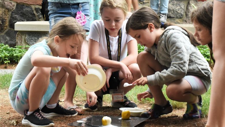 Three girls lean over a food-based science experiment outdoors at Penn State Behrend.