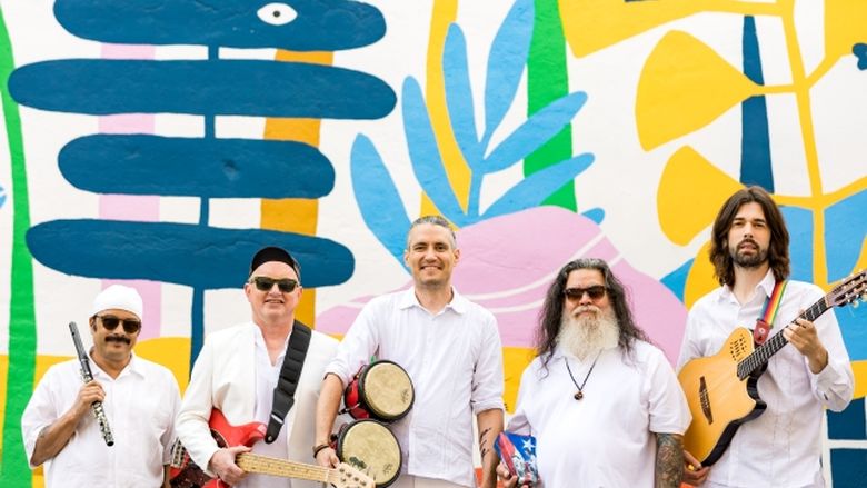 Publicity Photo: De Tierra Caliente band members standing with instruments against a vibrant mural.