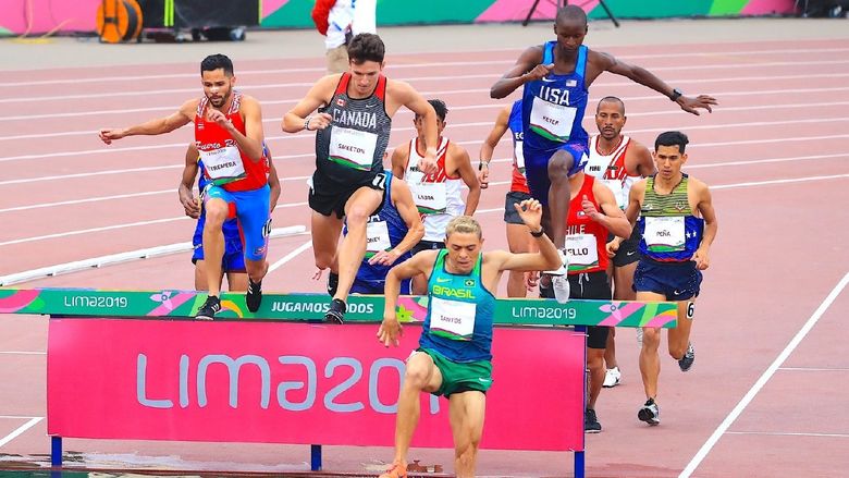 Runners jump over a hurdle and into water during a steeplechase race.