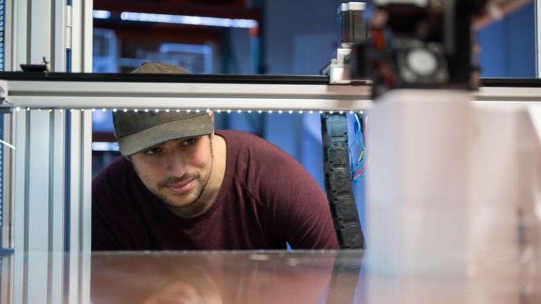 A student bends down to look into the printing bed of a 3D printer at Penn State's Innovation Commons.