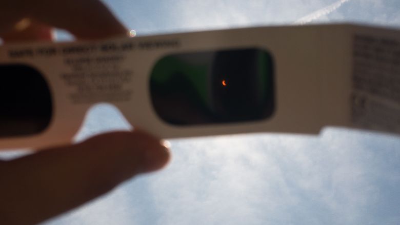 An image of a hand holding a pair of paper glasses for viewing the solar eclipse up to the blue sky