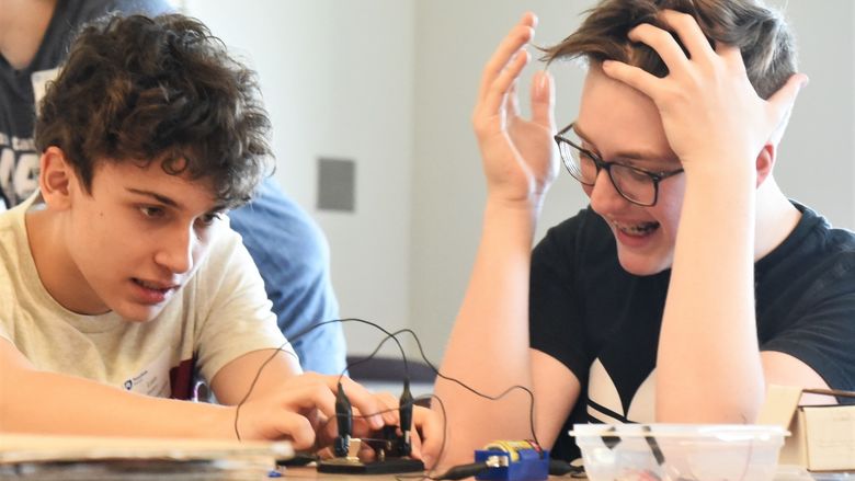A boy laughs and holds his hands to his head as another boy sends him a message using Morse code.