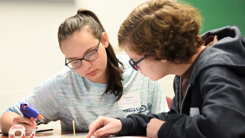 Two students build a model bridge using a glue gun and spaghetti noodles.