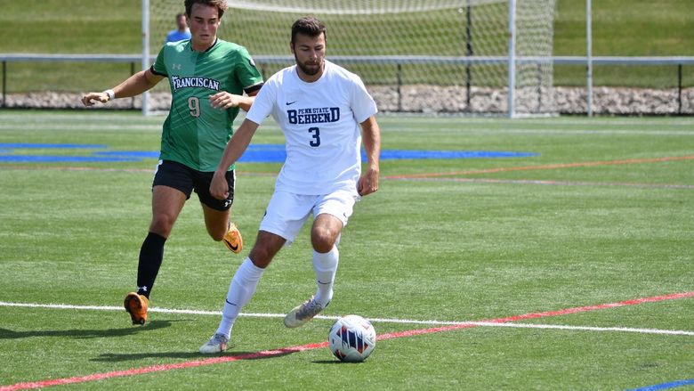 A Penn State Behrend men's soccer player advances the ball to midfield while being pressured by a defender.