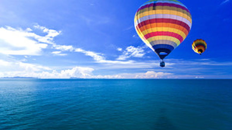 A colorful hot air balloon floats above blue water against a blue sky.