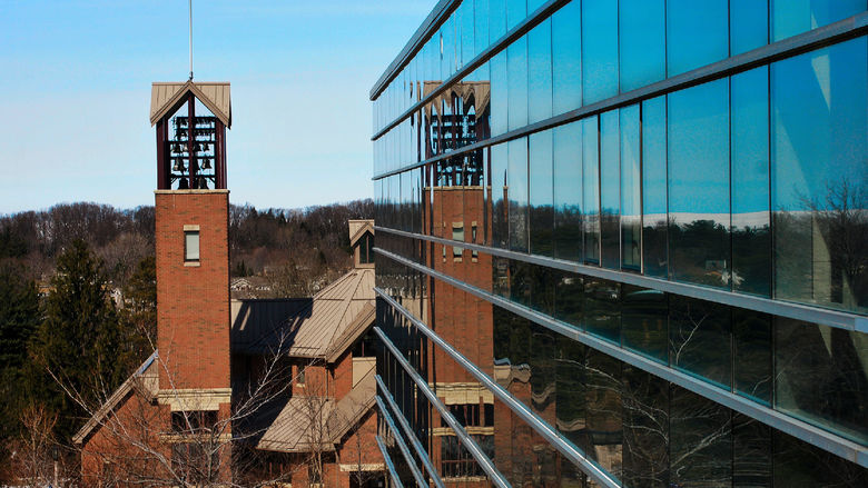 The Smith Carillon, a tall bell tower of red brick, as viewed from a building sitting beside it. The tower is reflected in the glass windows.
