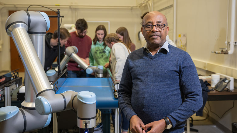 A professor stands in the Prischak Robotics and Automation Lab, with several students in the background.