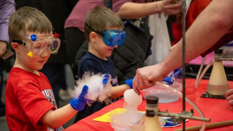 A boy wearing safety goggles holds out his hand during a chemistry demonstration.