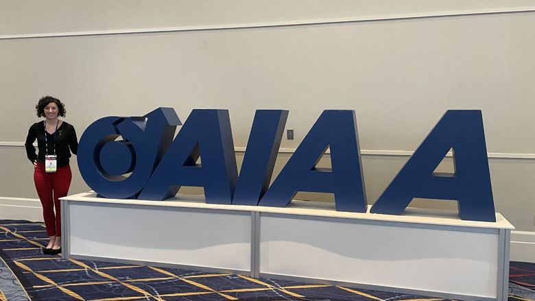 Renee Dorer stands next to the logo for the American Institute of Aeronautics and Astronautics, or AIAA