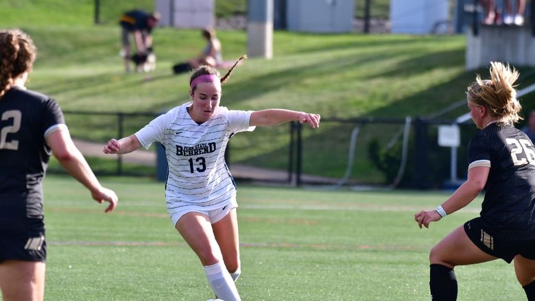 A Penn State Behrend soccer player dribbles between two defenders.