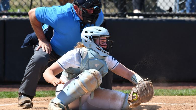 A catcher for the Penn State Behrend softball team grabs the ball as the umpire leans over her shoulder.