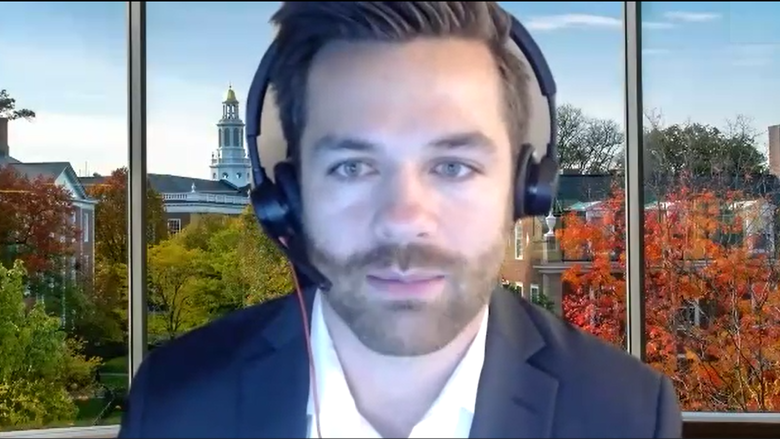 Tim Kluska, wearing headphones, gives a speech from his computer, wearing a suit and tie with the campus of Harvard in the background