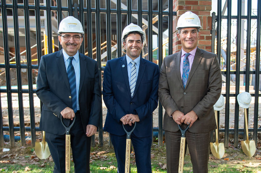 Three men wearing suits and hard hats pose with shovels in front of a blank fence.