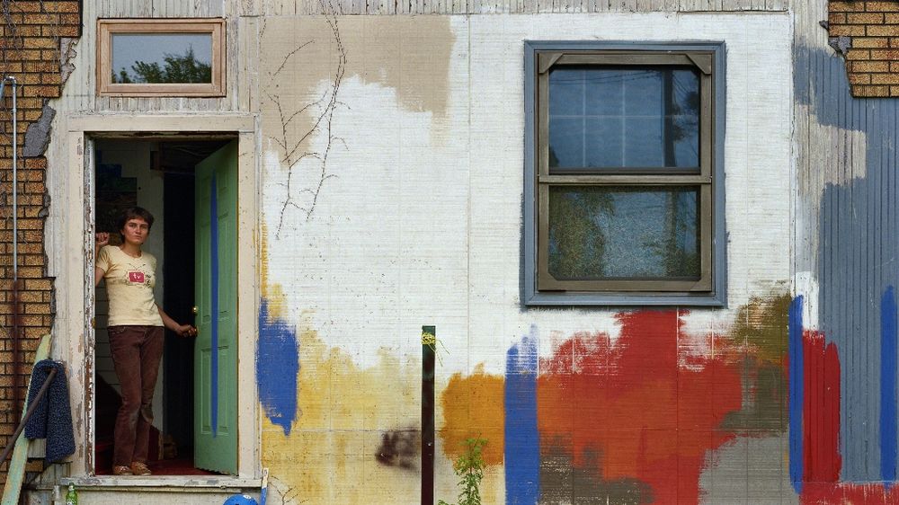 A woman stands in the doorway of a dilapidated house.