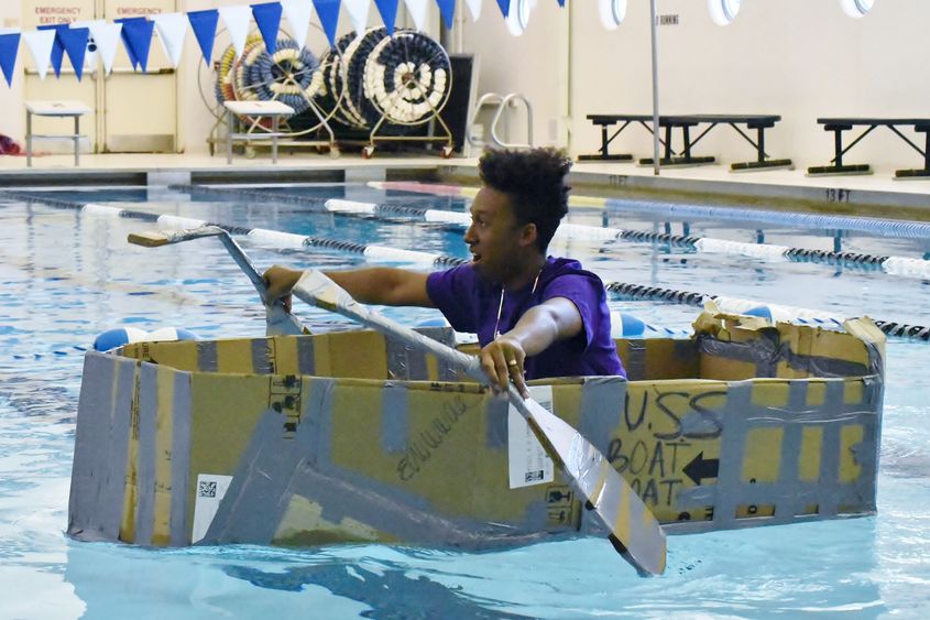 A high school student sails a cardboard boat in a swimming pool.