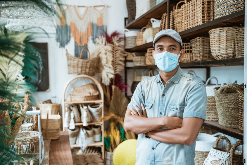 An employee in a hat and facemask poses in a shop stocked with wicker baskets.