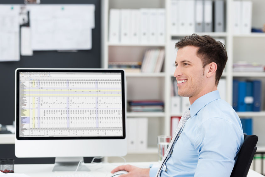 A young male office worker smiles while looking away from the spreadsheet on his computer.