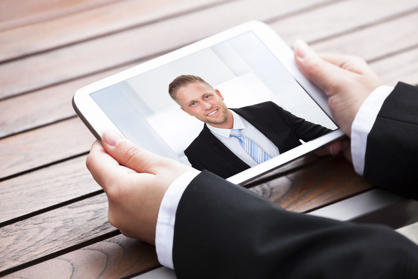 A man in a suit holds a tablet computer that shows another man in a suit.