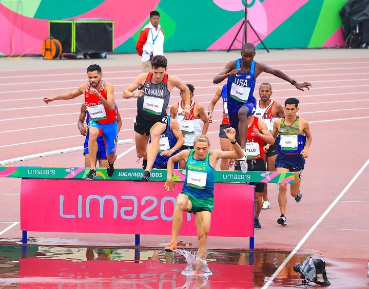 Runners jump over a hurdle and into water during a steeplechase race.